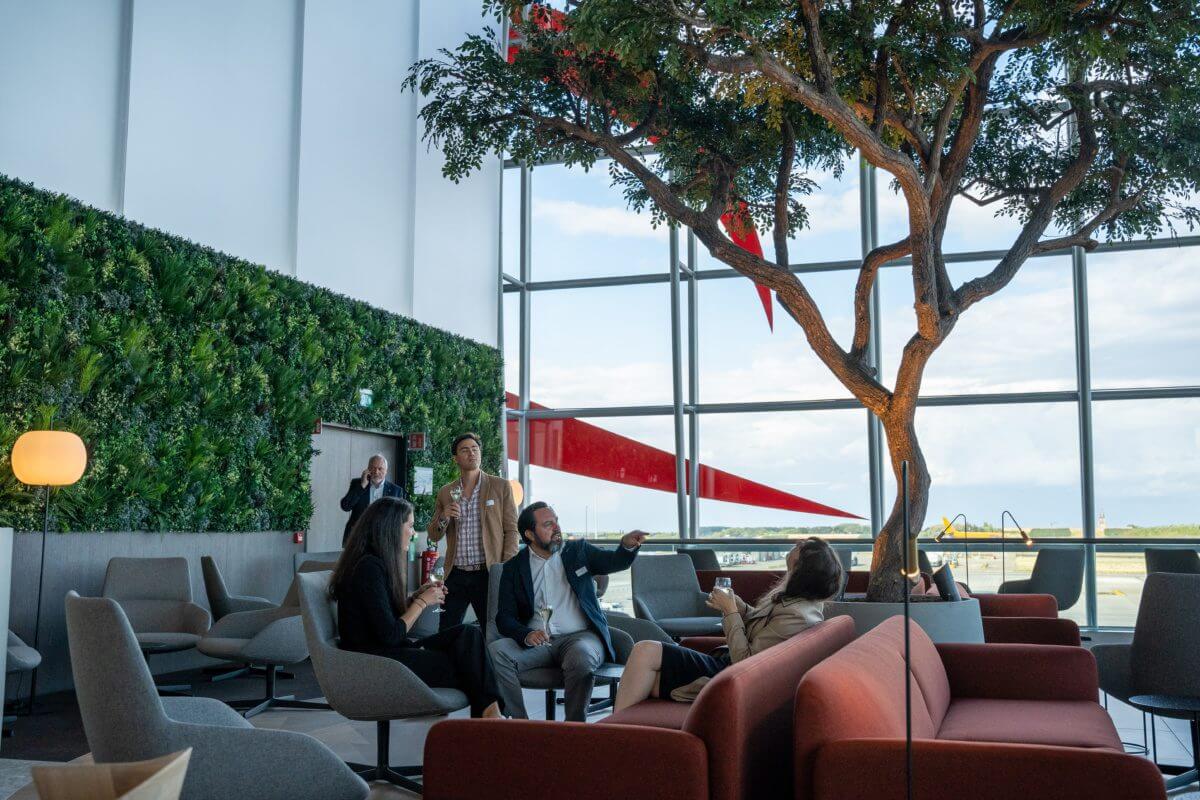 The Sunrise Lounge Green Wall at Brussels Airport offers a stylish setting with a green wall and large windows. In this modern space, several people in business attire are seated on sofas, conversing and relaxing beneath a large indoor tree that adds a natural touch to Belgium's finest lounge experience.