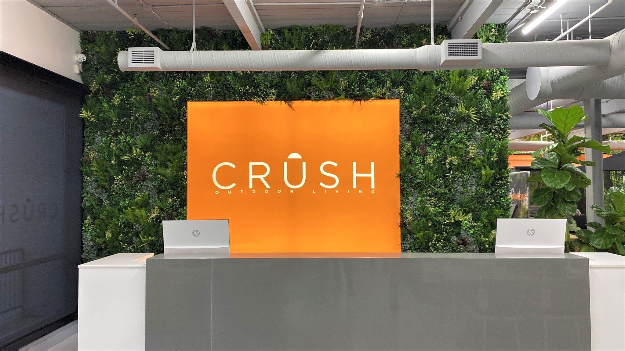 Reception area with a modern, minimalist design. A bright orange sign on a wall featuring a faux green wall reads CRÜSH OUTDOOR LIVING. Two white desks with computers are in the forefront, and there is a large potted plant to the right, capturing the essence of this showroom in Toronto, Canada.