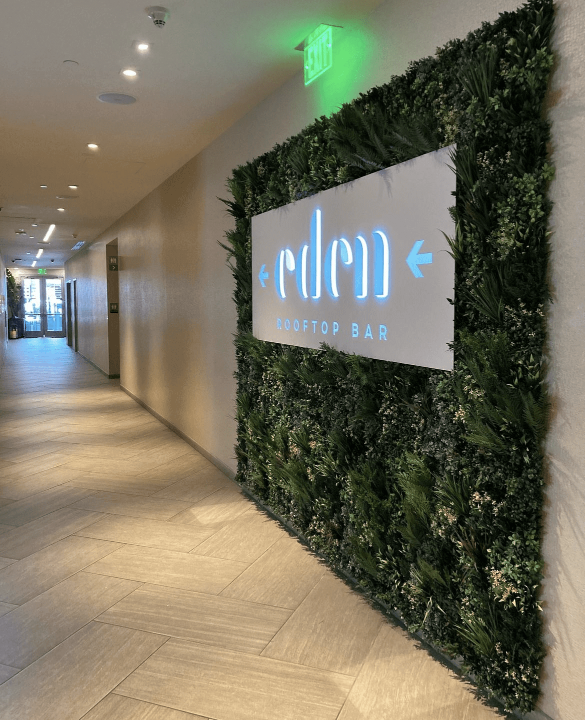 Discover Eden Rooftop Bar in Phoenix, Arizona, where neon lighting illuminates a lush wall of green foliage. The well-lit corridor, marked by an exit sign above, guides you to the inviting door ahead.