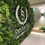 Wall with lush greenery, mimicking a faux green wall in a Health Spa in Stockholm, Sweden, featuring a logo that includes the letters S and V encircled by laurel leaves. Below the logo, the text reads SVERIGES SKÖNHETS CENTER in white, accompanied by a soft light on the right.