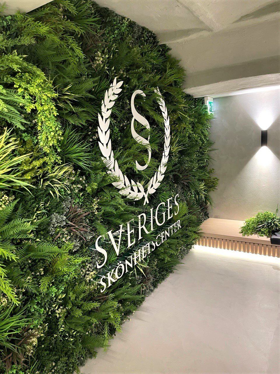 A faux green wall at a health spa in Stockholm, Sweden, displays the text Sveriges Skönhetscenter in white letters. The lush variety of plants is complemented by a logo featuring a stylized letter S encircled by a laurel wreath.