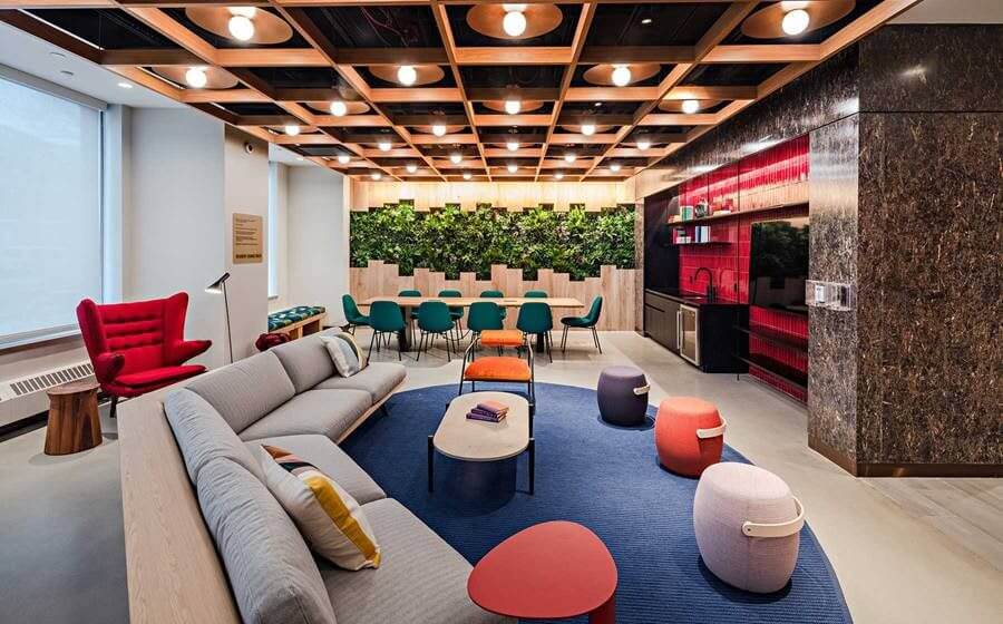 A modern, brightly lit lounge in a multi-family facility in Brooklyn features a gray sectional sofa, colorful round stools, and a blue rug. In the background, a dining area with green chairs sits against a lush vertical garden. The ceiling boasts a geometric wooden design with circular lights.