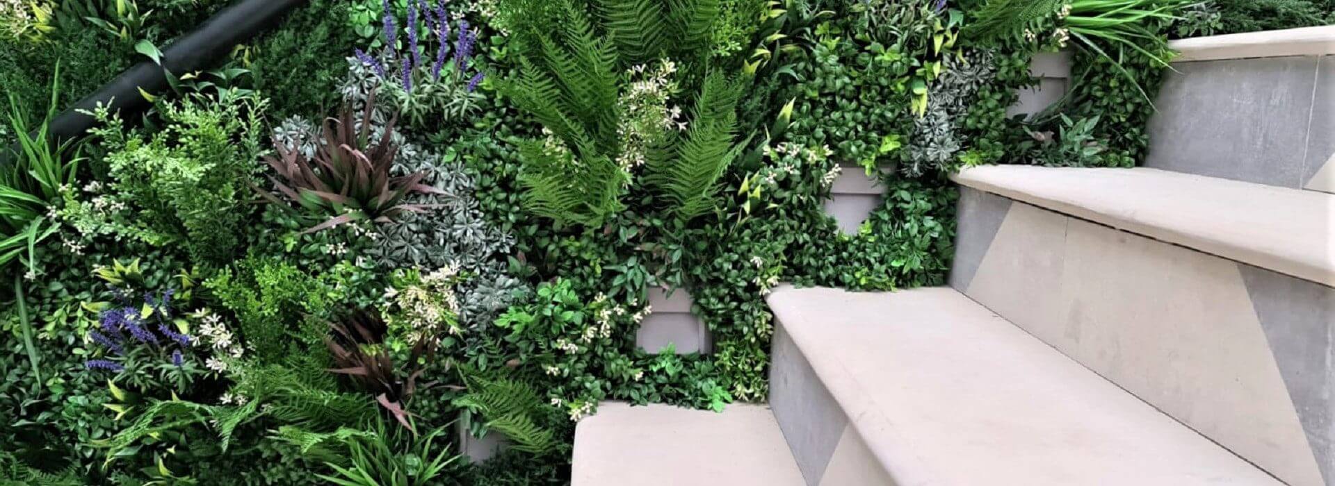 A staircase with light-colored steps is adjacent to a lush vertical garden in London, UK. The stairway green wall is filled with various plants, including ferns and small purple flowers, creating a vibrant and natural backdrop.