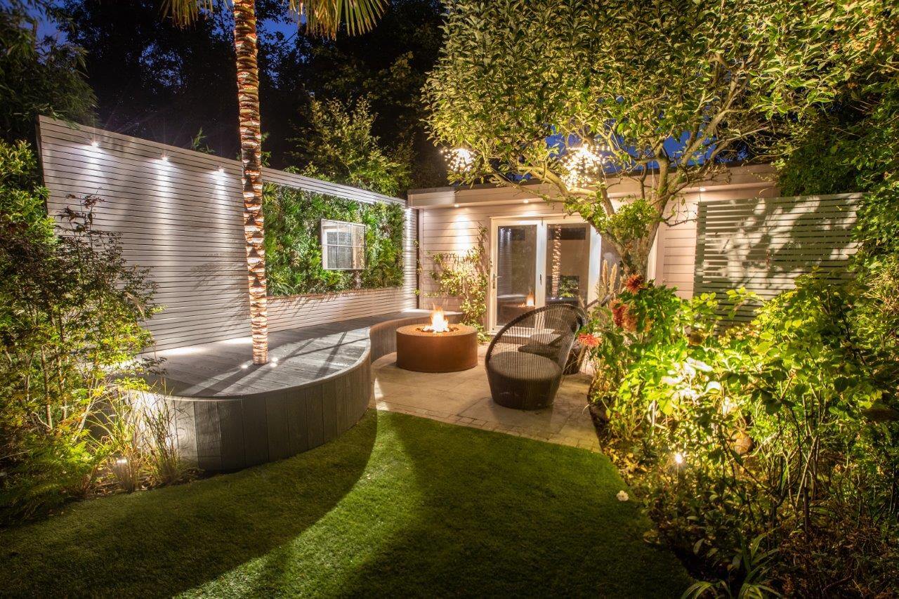 A cozy backyard at night with warm lighting, featuring a fire pit surrounded by seating, lush green plants, and a small deck adorned with an artificial green wall. The scene conveys a tranquil, inviting atmosphere, perfect for inspiration.