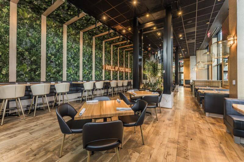 A modern restaurant interior with wooden floors and tables is reminiscent of Moxies Restaurant in Winnipeg. Black and white chairs are arranged neatly, while a lush green wall features a neon sign. Tall windows and elegant lighting create an inviting atmosphere, reflecting the charm of Manitoba.