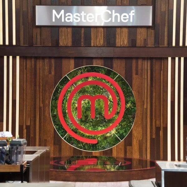 A kitchen set for a cooking show features a wooden backdrop with an artificial green leafy circle displaying a red "m" logo. The word MasterChef is visible at the top, with kitchen counters and cooking equipment in the foreground.