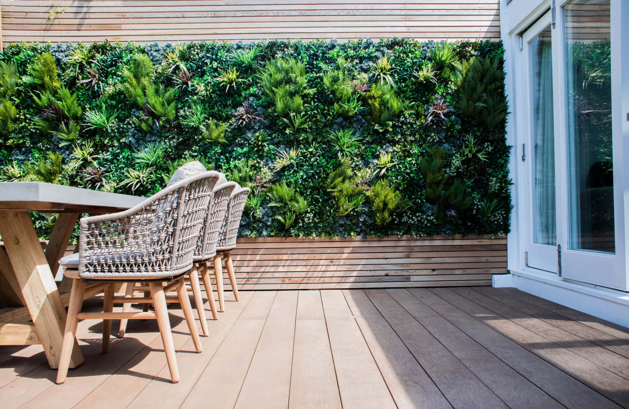 A modern outdoor patio with a wooden deck, featuring a dining table with wicker chairs. A vertical garden with lush green plants covers the wall, providing a natural backdrop. Glass doors are partially visible on the right.