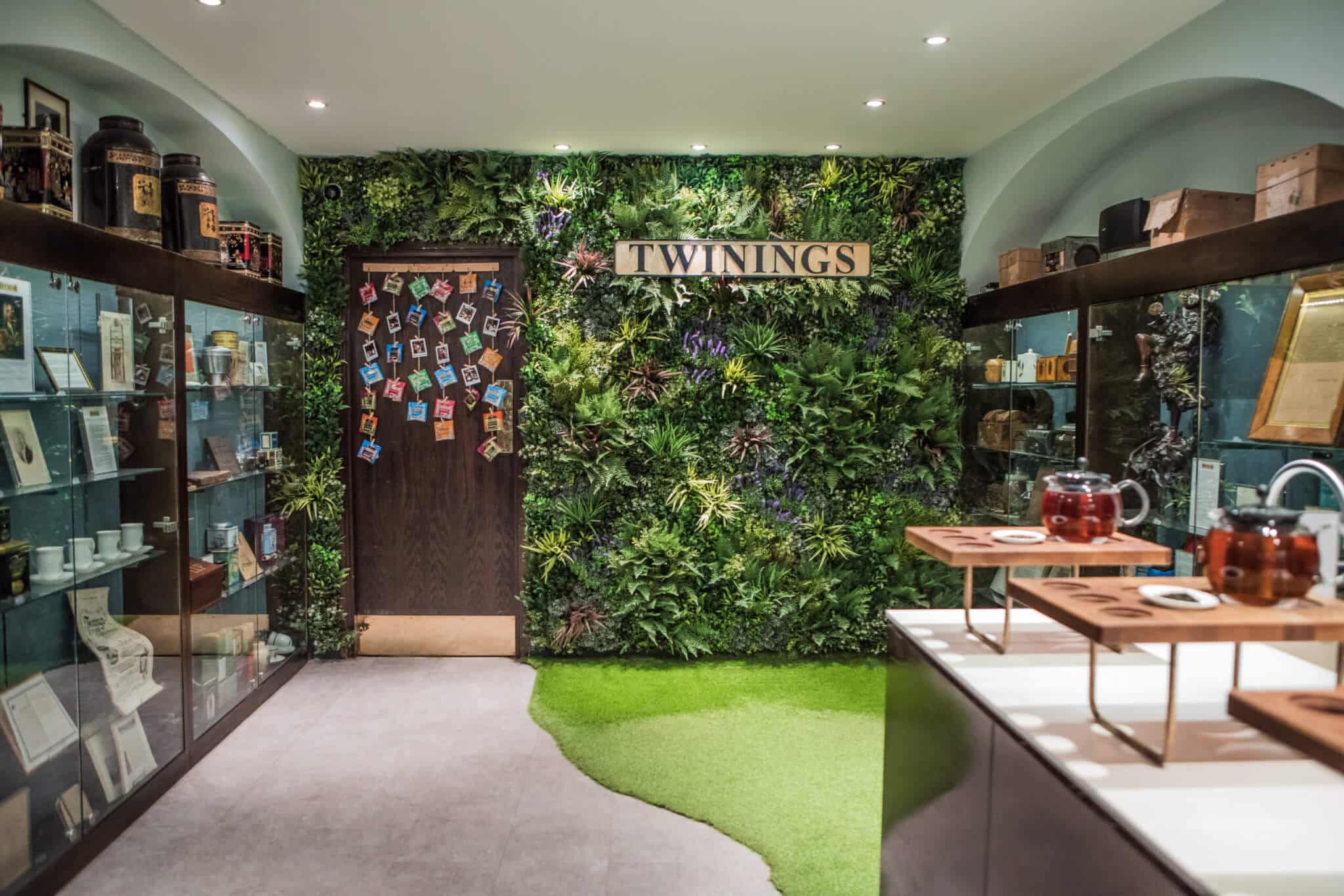 A cozy tea room with a lush green wall, displaying the Twinings sign. Shelves on either side hold tea items and memorabilia. A door adorned with colorful cards adds charm. Tables exhibit teapots, enhancing the serene atmosphere.