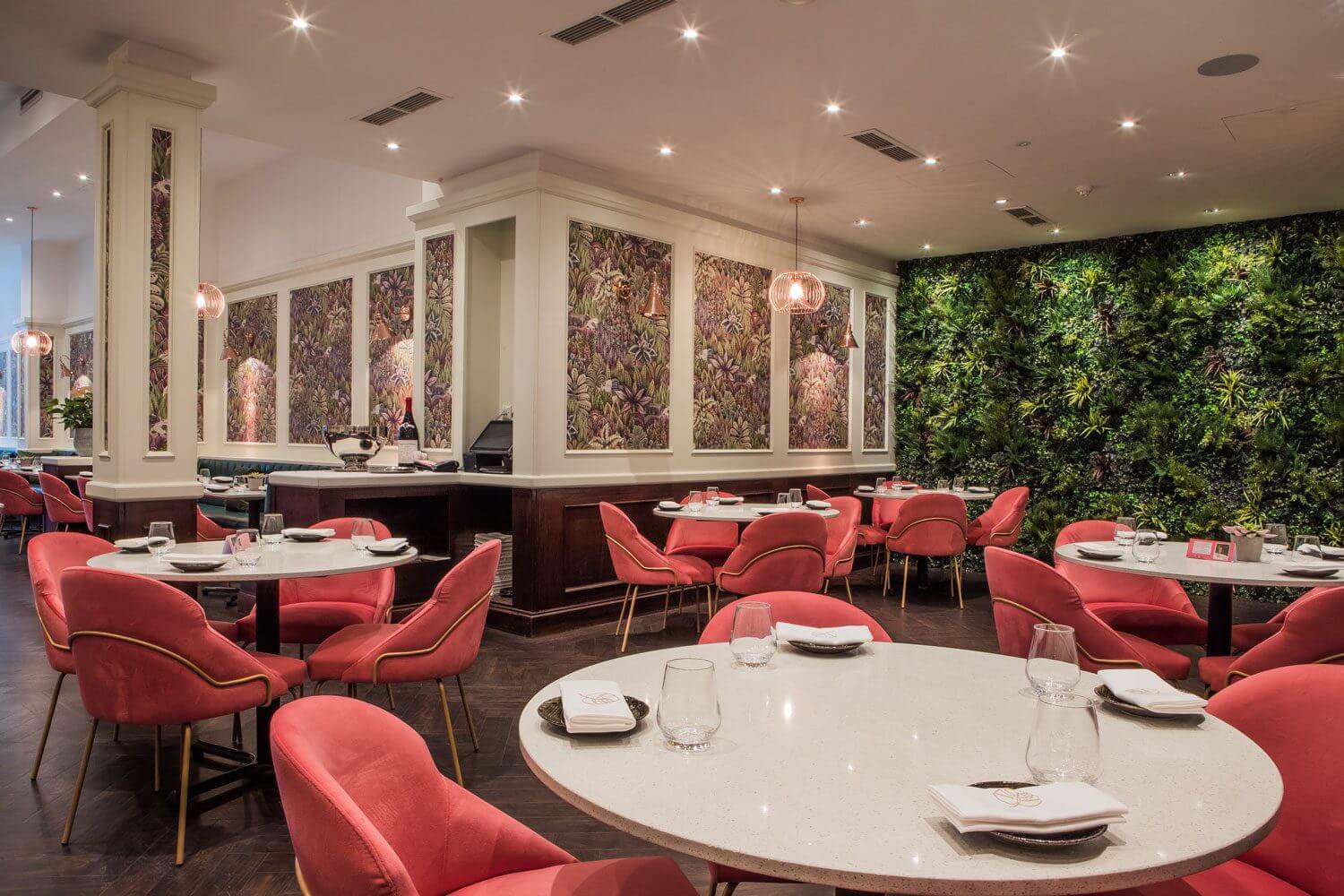 A modern restaurant interior with round tables and red upholstered chairs. The walls feature decorative panels and a vertical garden. Soft lighting from ceiling fixtures creates a warm ambiance. Tables are set with glasses and folded napkins.