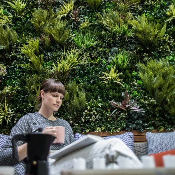 A person sits on a couch holding a pink mug, with a lush green vertical garden wall in the background. A black coffee pot is on the table in the foreground. The setting evokes a cozy, artificial yet natural atmosphere.