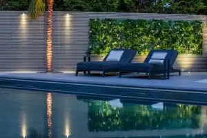 Two black lounge chairs with cushions are positioned beside a calm, illuminated pool. A vertical garden and a palm tree are in the background, enhanced by artificial ambient lighting for a serene atmosphere.