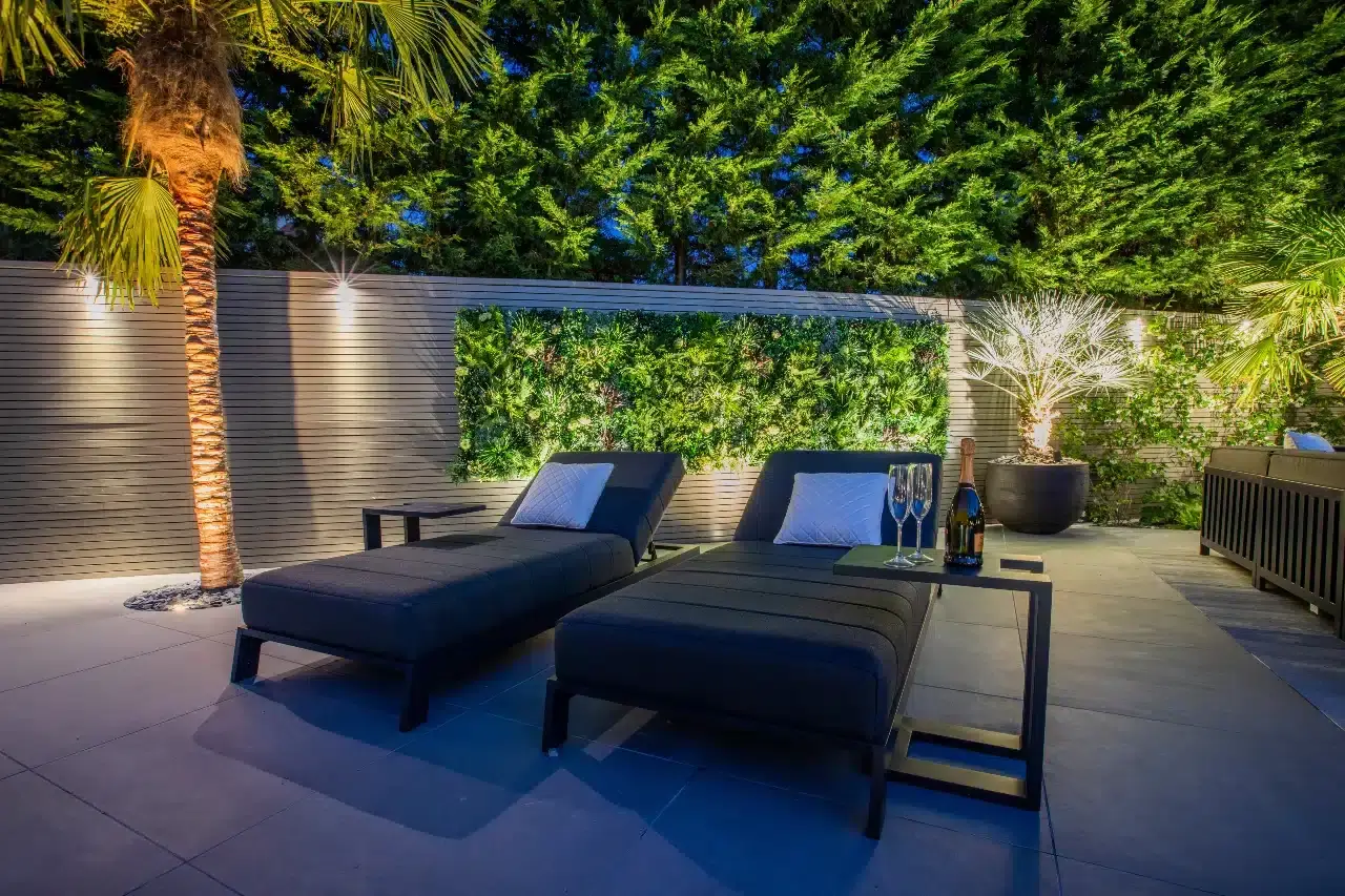 Modern outdoor patio with two dark loungers, a small table with glasses, surrounded by lush greenery. A tall palm and a wall with vertical garden add a tropical touch. Evening lighting creates a cozy ambiance.