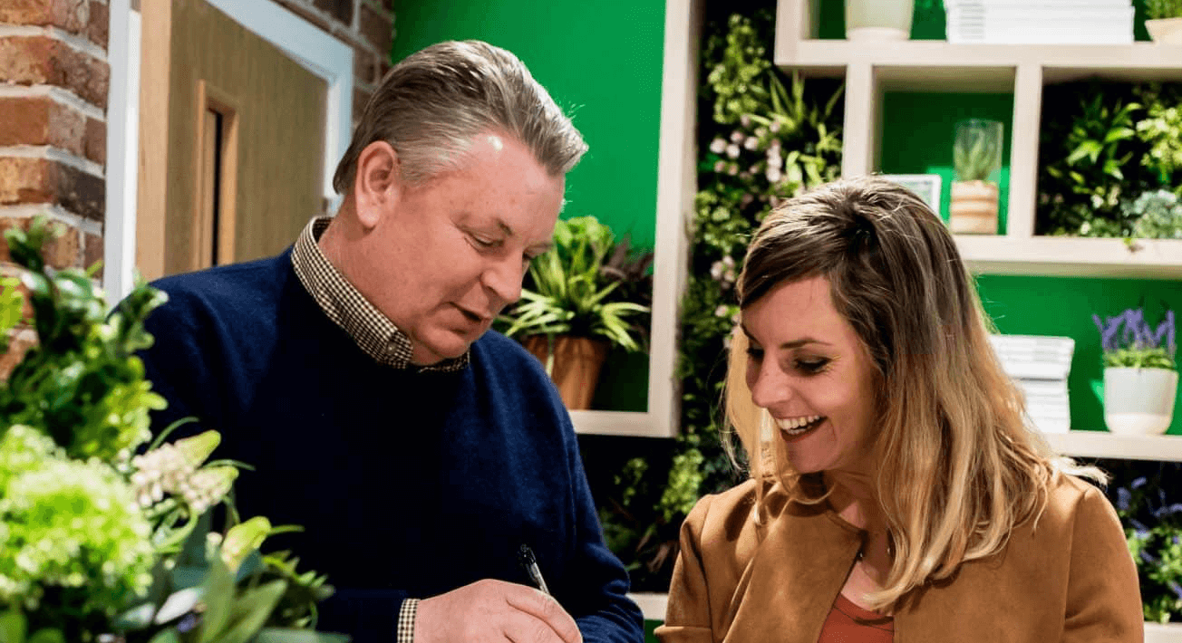 A man and a woman are in a room decorated with plants and shelves. The man, holding a pen, is looking at papers while the woman smiles, appearing engaged in conversation. The setting suggests a casual, friendly meeting or discussion.