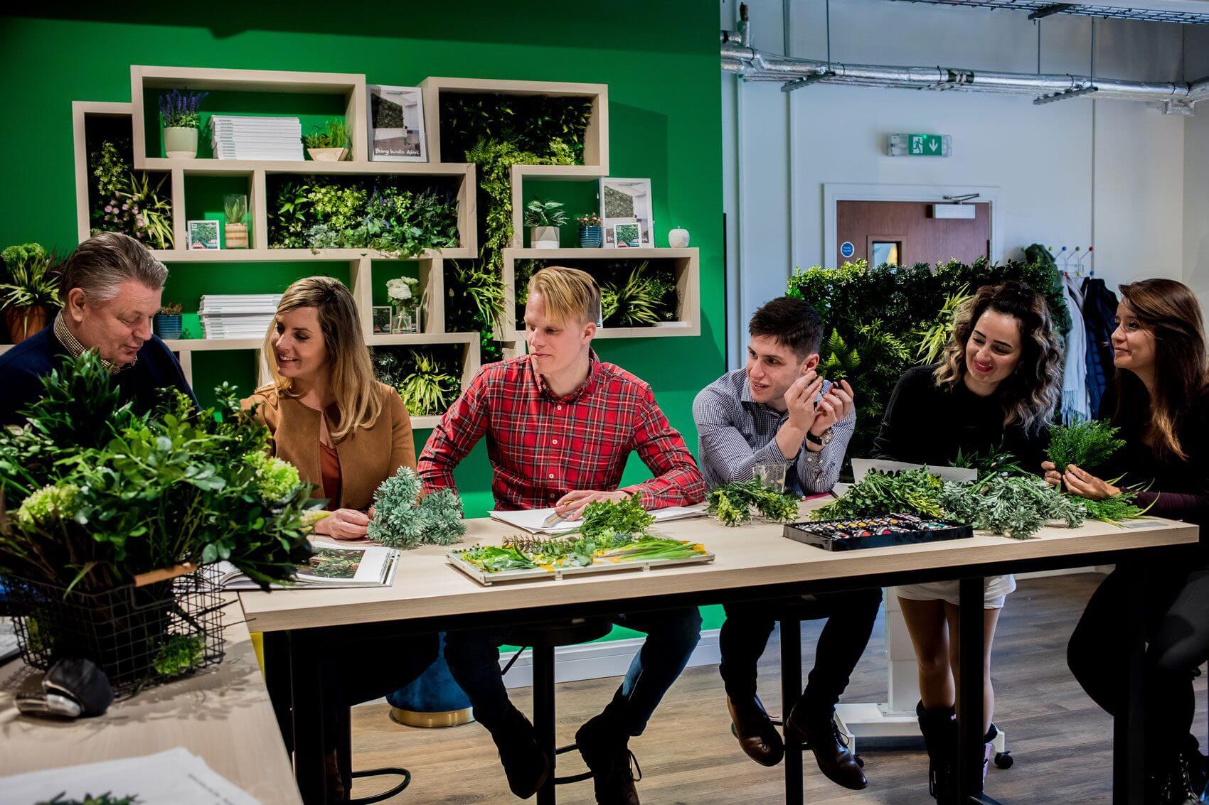 A group of six people, three men and three women, are sitting and standing around a table covered with various green plants. They appear engaged in discussion, surrounded by green walls and shelves holding plants.