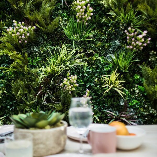 A lush vertical garden featuring artificial green walls comes alive with various plants and small pink flowers. In the foreground, there's a table with a glass of water, a potted succulent, and a bowl of oranges.