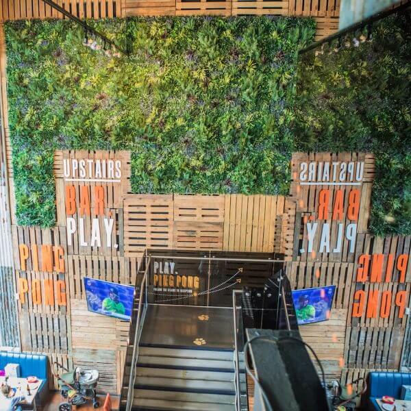 A vibrant interior with a vertical garden wall, part of the Green Walls Retail Concept in Stratford, and signage reading UPSTAIRS BAR PLAY PING PONG. A staircase leads upward while two screens display sports, overlooking a cozy seating area below.