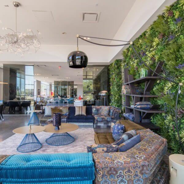 A modern lounge area with colorful patterned sofas and blue accent pillows. An artificial vertical garden covers one wall, and round coffee tables with artistic wireframe lamps are in the center. Large windows allow natural light to fill the space.