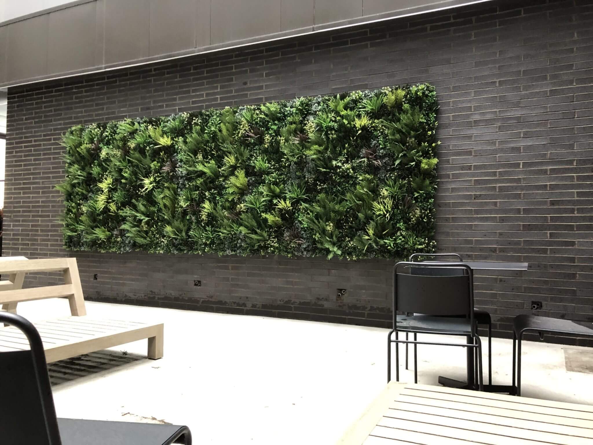 A modern outdoor patio with a vertical garden on a brick wall complements the Artificial Roof Garden Project in Manchester, UK. The area features wooden benches and a dining table with metal chairs, crafting a relaxed and natural ambiance.