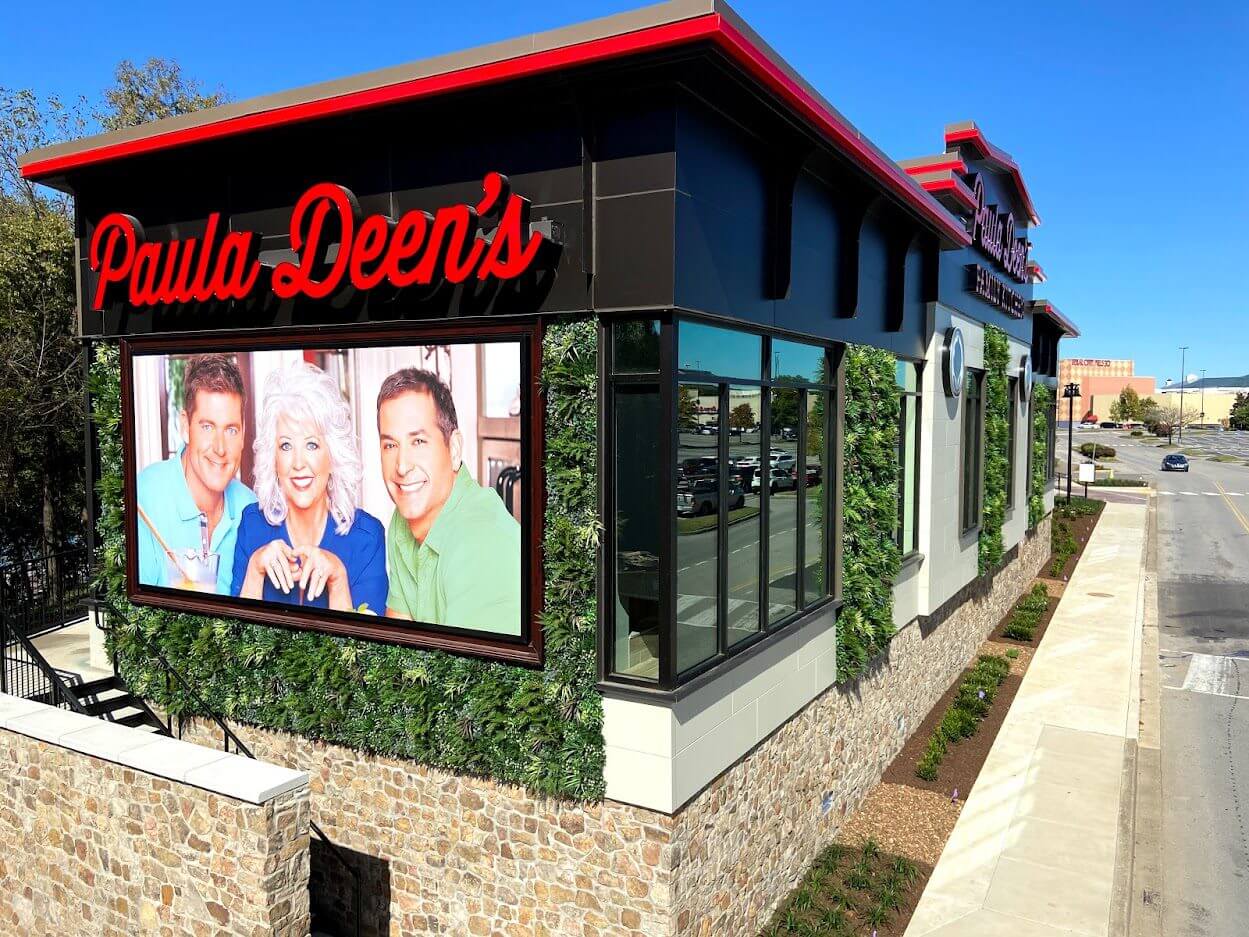 The image shows an exterior view of Paula Deen's Restaurant in Nashville, Tennessee, with a faux green wall enhancing the facade. A billboard with three smiling individuals adorns the side. The building is surrounded by greenery under a clear blue sky.