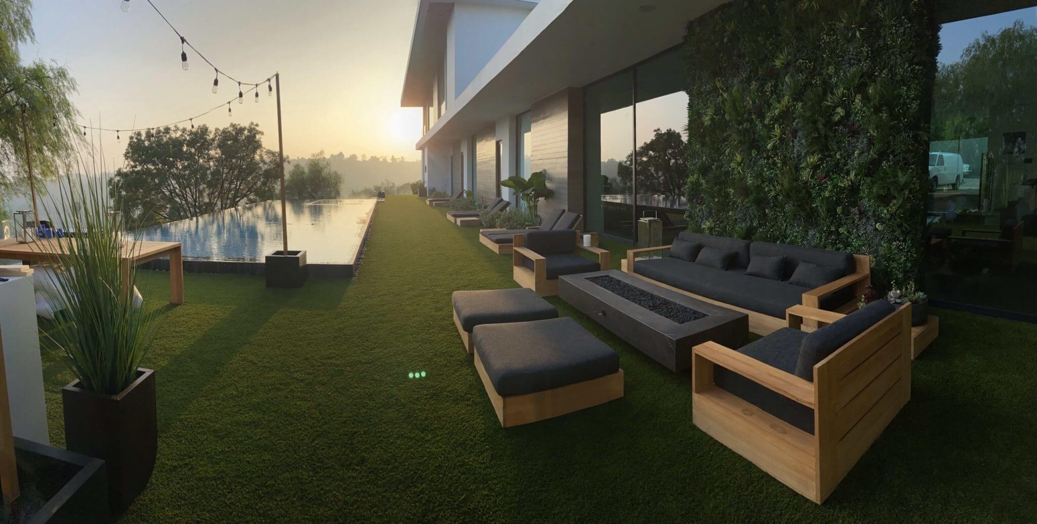 At sunrise, a modern outdoor patio features wooden furniture, lush green grass, and a pool reflecting the sky. String lights dangle above, casting a warm glow, with an artificial vertical garden on the wall of a sleek building. Trees stand gracefully in the background.