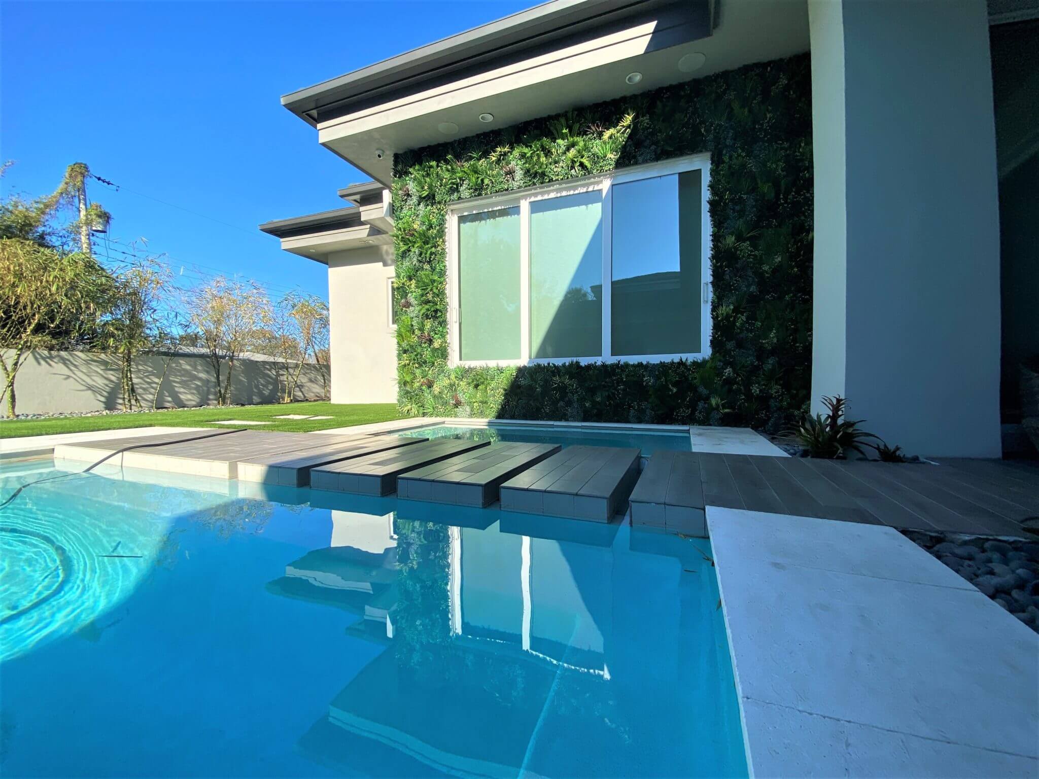 A modern house in Denver boasts large windows and a green wall that merges seamlessly with a rooftop garden. In the foreground, a swimming pool with a wooden deck complements the small garden detail, all set against Colorado's clear blue sky.