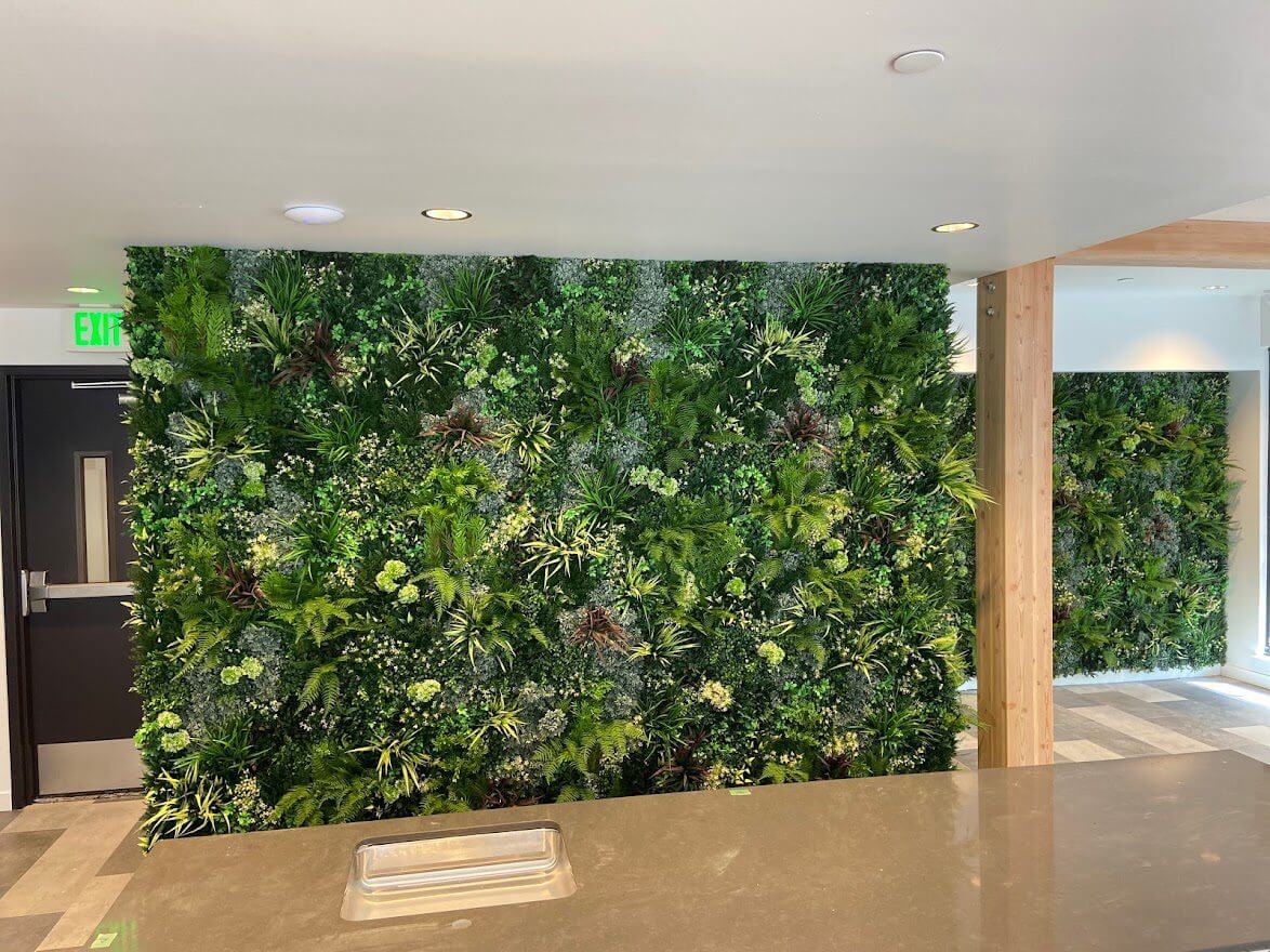 A modern indoor space in a Seattle multi-family residence features a large, lush vertical garden wall covered in faux plants and foliage. The room boasts wooden accents and recessed lighting, creating a fresh and calming atmosphere reminiscent of Washington's natural beauty.