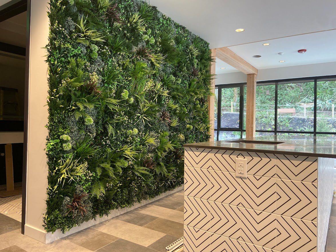 A modern interior features a large artificial green plant wall on the left and a reception desk with geometric patterns on the right. Sunlight streams through large windows in the background, creating an inviting atmosphere.