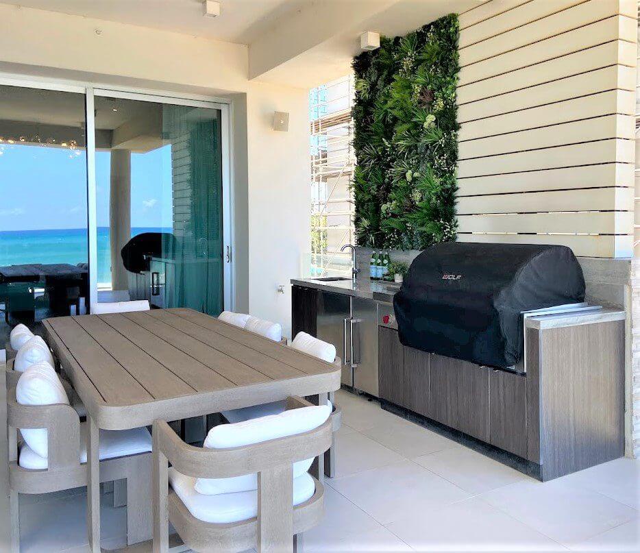 Outdoor terrace in a private residence with a modern dining set and built-in grill. The space features a wooden table with cushioned chairs, a grill covered with black cloth, and a lush green wall. In the background, there's an ocean view and sliding glass doors, capturing Puerto Rico's beachside charm.