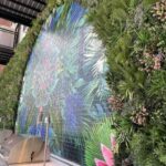 A vibrant green wall of artificial plants surrounds a large mural with colorful floral patterns on a brick surface. Below, two stainless steel barbecue grills are positioned on a countertop. An industrial-style balcony is visible above.