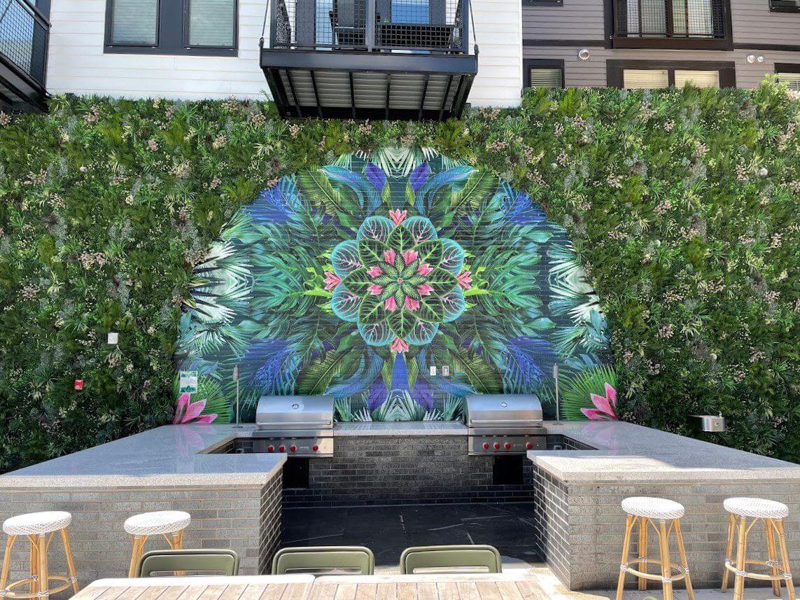 An outdoor grill area with two grills set into a stone counter boasts a vibrant mural of green leaves and pink flowers. The lush vertical garden backdrop integrates an artificial green wall, enhancing the ambiance in this multi-family complex. Bar stools are placed in front of the counter.