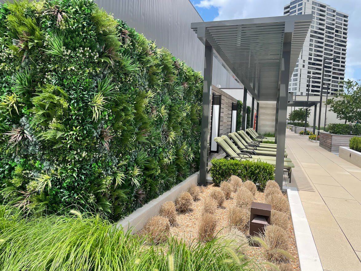 A modern rooftop garden with a vertical green wall adorns the Fitness Center in Houston, Texas. Lounge chairs rest under a pergola, while ornamental grasses sway in the foreground, framed by a tall building visible in the background.