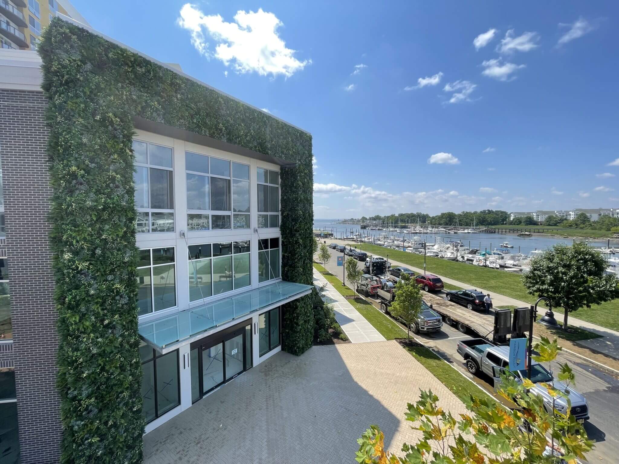 A modern building with an external facade green wall in Stamford stands next to a marina filled with boats. The sky over Connecticut is clear and blue, with a few clouds. A tree- and grass-lined sidewalk runs alongside a road dotted with parked vehicles.