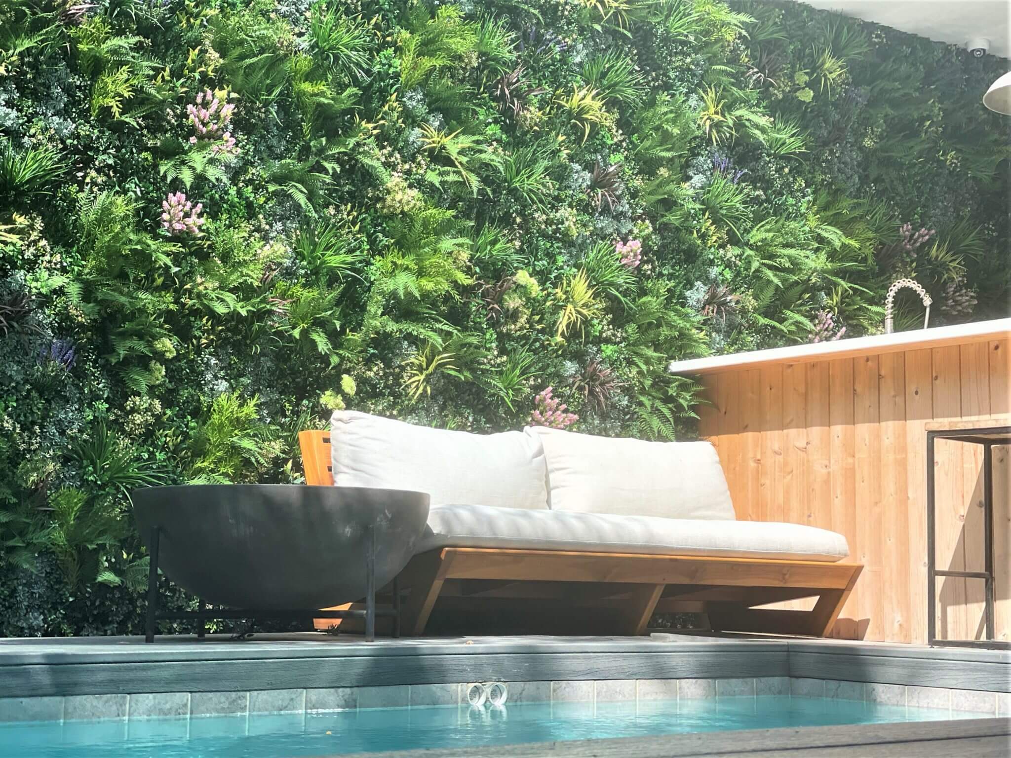 A cozy outdoor lounge area showcases a wooden bench with white cushions next to a small pool. The space is enhanced by a faux green wall installation and features a wooden bar counter with a modern sink, creating an inviting poolside retreat.