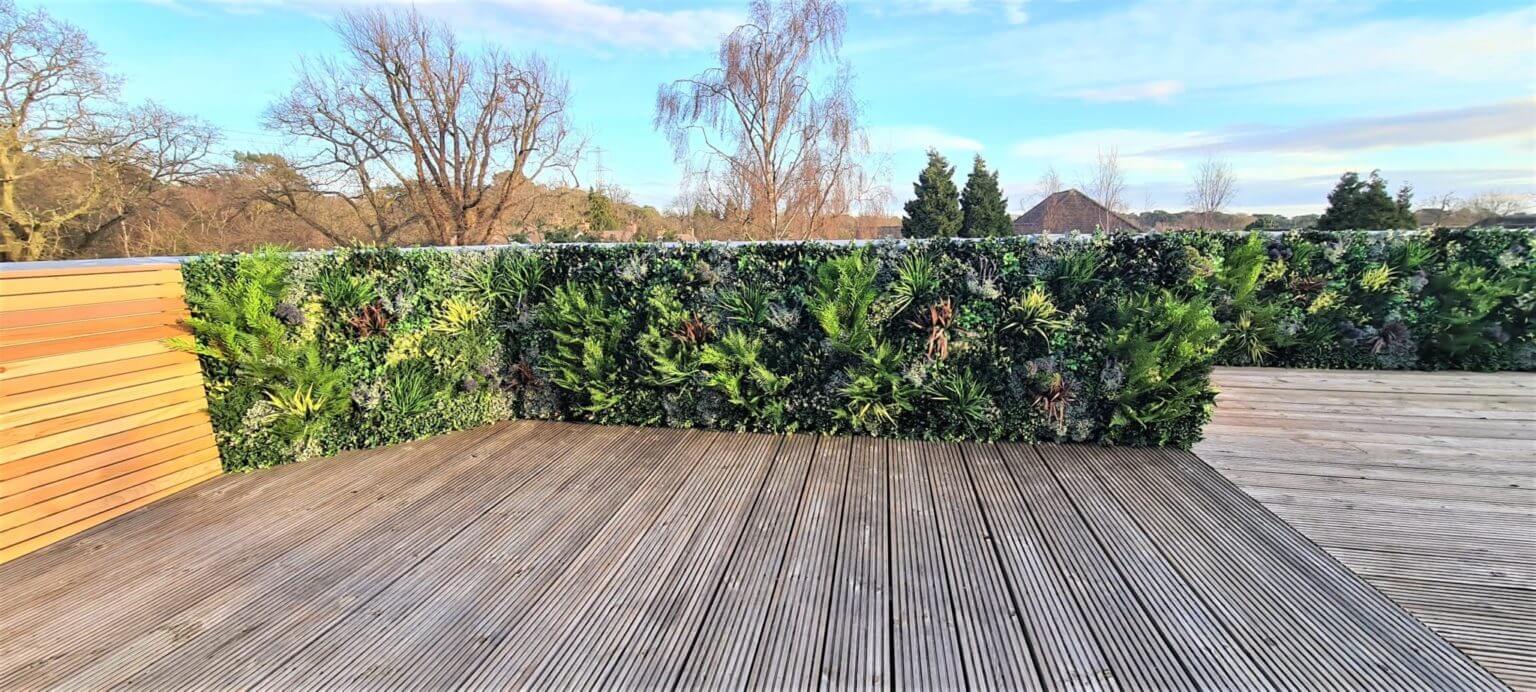 A rooftop terrace with wooden decking and walls covered in lush, artificial green foliage. A clear sky and leafless trees are visible in the background, suggesting a winter or early spring setting.