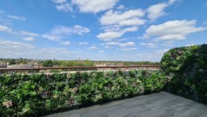 A rooftop terrace with an artificial green wall and a clear sky above overlooks a distant cityscape under a blue sky scattered with fluffy white clouds.