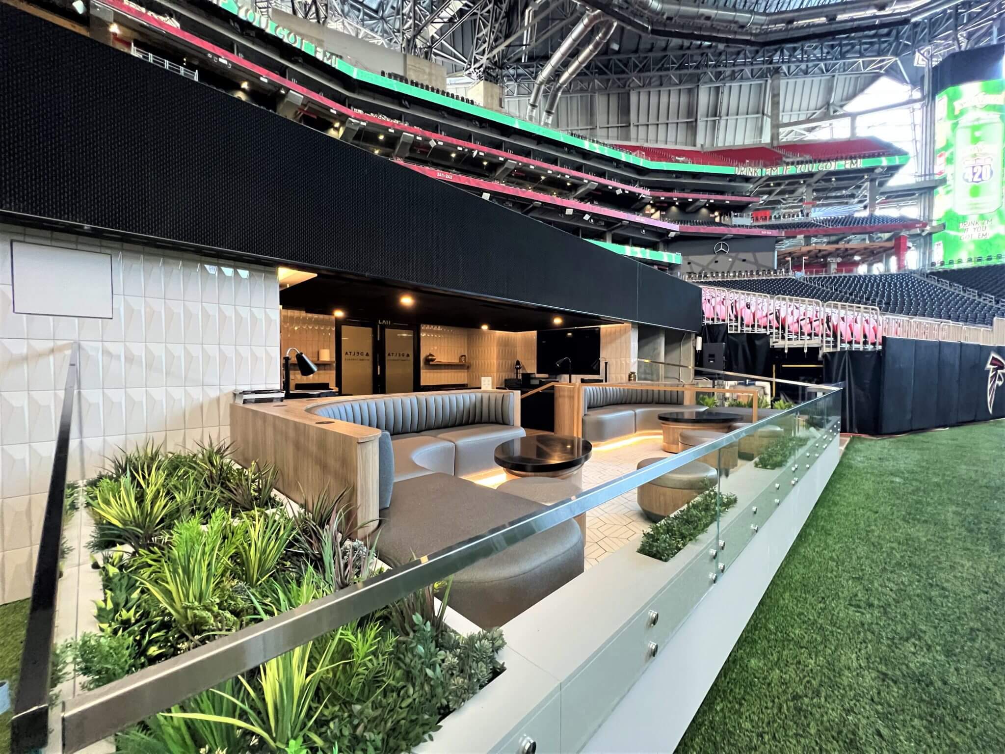 Luxurious stadium seating area with modern, cushioned booths and a glass barrier. Artificial plant installations decorate the space, overlooking the field at Mercedes Benz Stadium in Atlanta, Georgia. The open-air design offers views of the stadium roof and seating sections in the background.