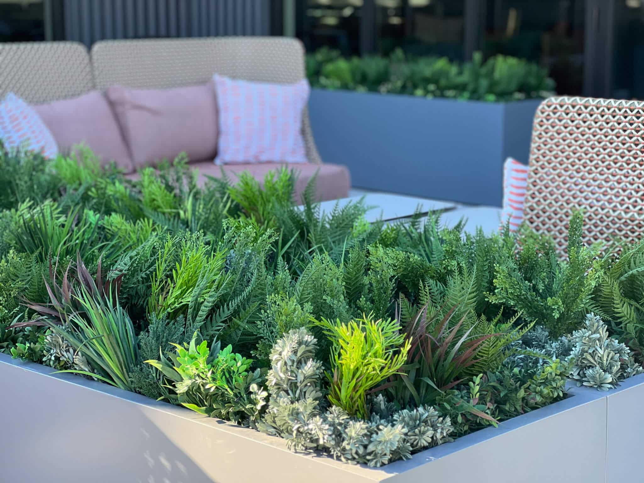 A cozy outdoor seating area featuring a large planter box filled with Loose Artificial Plants for Planters and Pots, showcasing a variety of lush green foliage and ferns. In the background, cushioned chairs and a sofa contribute to the relaxed atmosphere.