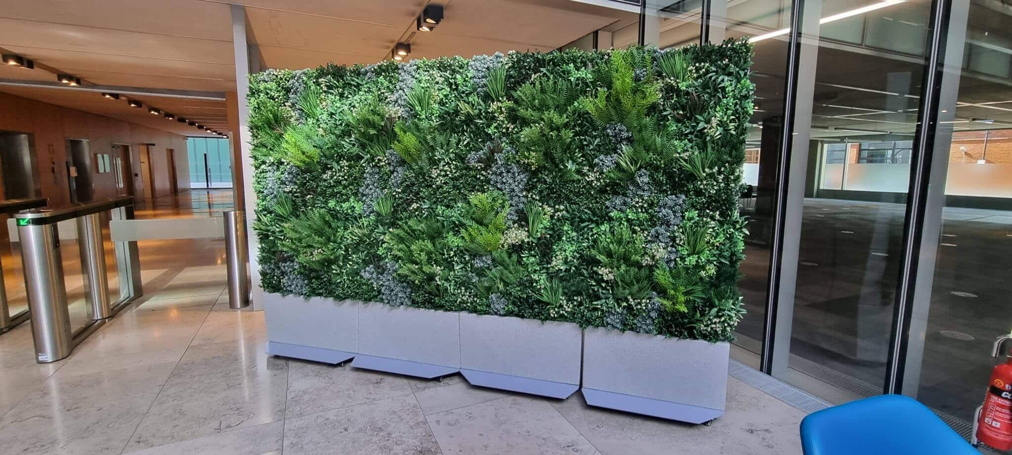 A wall inside an office building is adorned with lush artificial green plants, forming a vertical garden. The surrounding area features marble flooring, glass doors, and modern lighting, creating a sleek and natural ambiance. A blue chair is partially visible on the right.