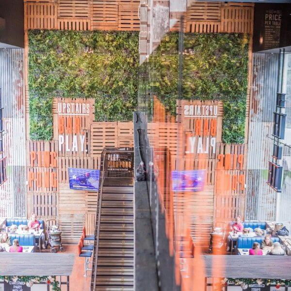 View of a modern urban space in Stratford, UK, where a green wall complements the large text reading UPSTAIRS BAR PLAY. Seating areas host people enjoying food, and the scene beautifully mirrors on a glass surface, embodying an innovative retail concept.