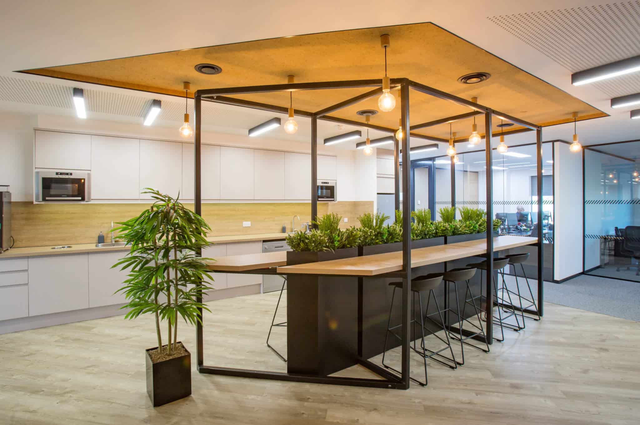 A modern office kitchen features a sleek island counter surrounded by black bar stools. Hanging lights illuminate the space, and the Luxury Flowering Panel adds a touch of greenery. White cabinets and appliances complete the contemporary look.