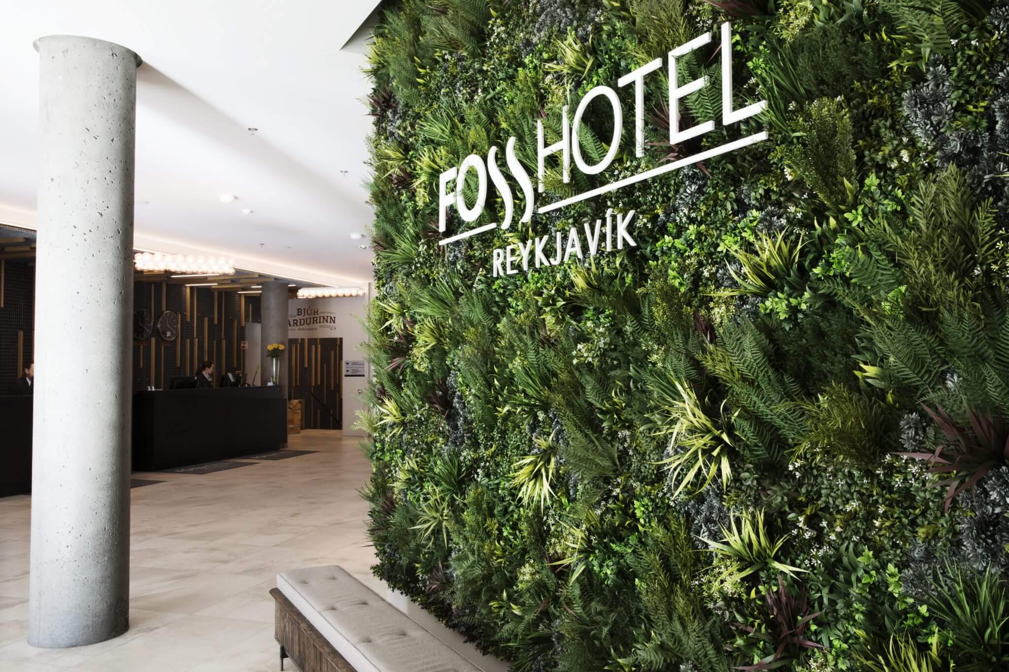 The entrance of Fosshotel Reykjavik showcases a stunning Hotel Green Wall in Reykjavik, with the hotel's name in sleek white letters. In the background, you can see the inviting reception area complemented by a concrete column and warm wood accents, reflecting Iceland's natural charm.