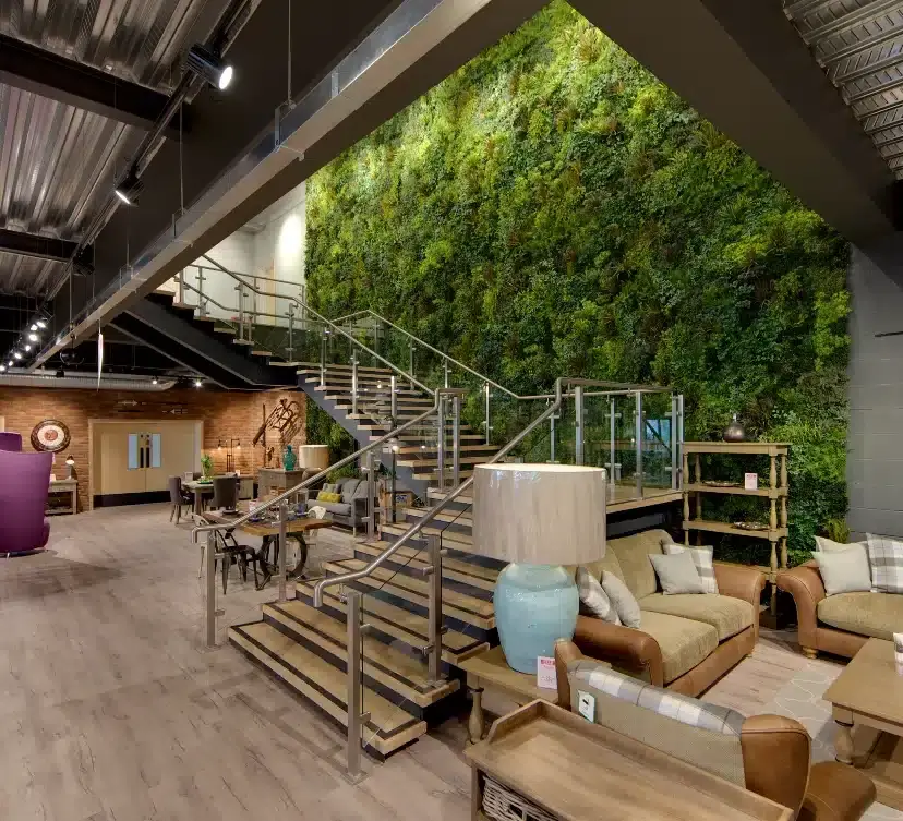 A modern interior showroom with a vertical green wall, a sleek metal staircase, and wooden floors. The space is furnished with contemporary sofas, tables, and lamps, creating a cozy and stylish atmosphere.