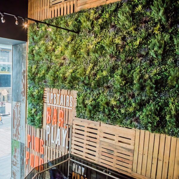 A vertical garden with lush, artificial green plants on a wall is combined with wooden panels displaying the words UPSTAIRS BAR PLAY. The scene is part of a modern indoor space, enhanced by string lights above.