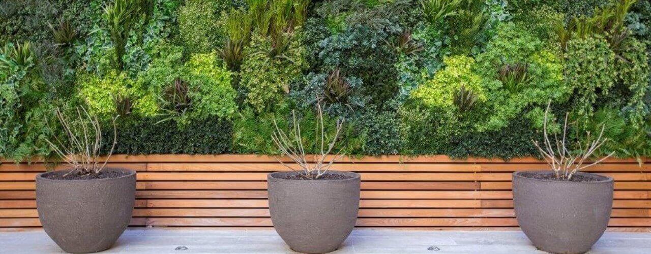 A vertical garden with lush green plants graces the wall. In front, three large, round, gray pots with bare branches sit on a wooden bench platform. The vibrant yet minimalistic scene subtly echoes the low-maintenance allure often associated with artificial greenery.