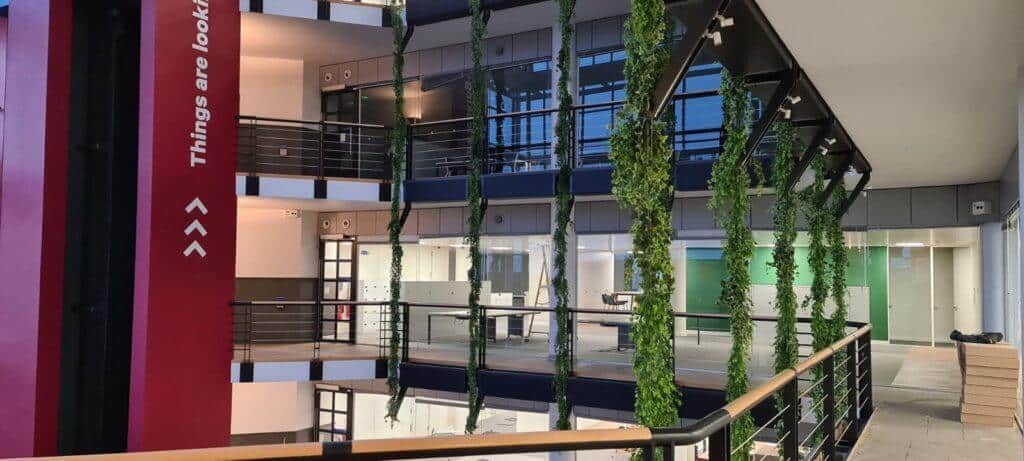 Modern interior with open floors, featuring a red column with text, glass railing, and artificial green plants hanging from the ceiling. Offices with glass walls are visible on each side, creating a bright and spacious atmosphere.