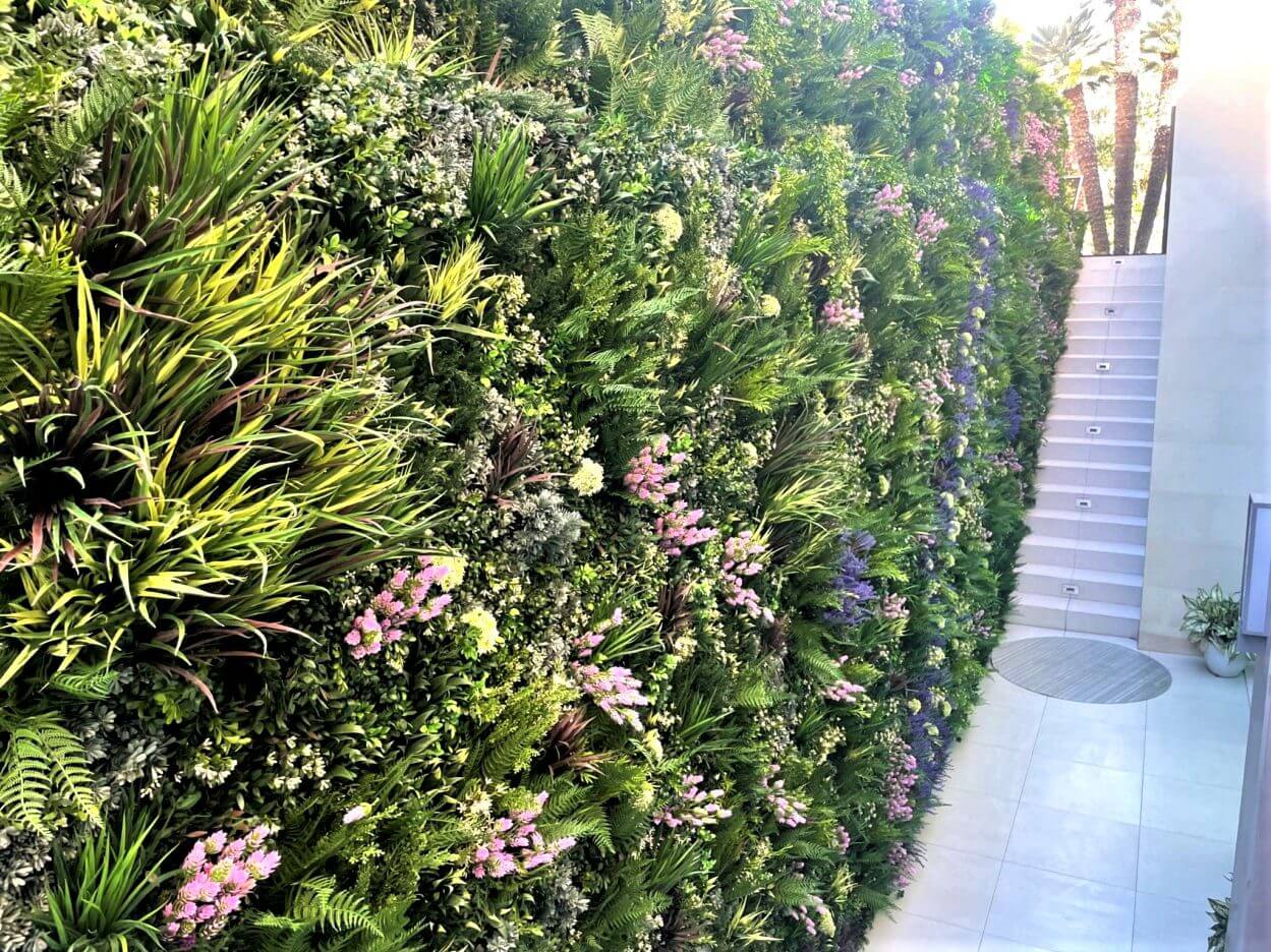 A bespoke designed green wall in Laguna Beach, California features a lush vertical garden with diverse green foliage and clusters of pink and purple flowers lining the wall. Adjacent to a white-tiled staircase, potted plants at the base enhance this well-lit outdoor oasis.