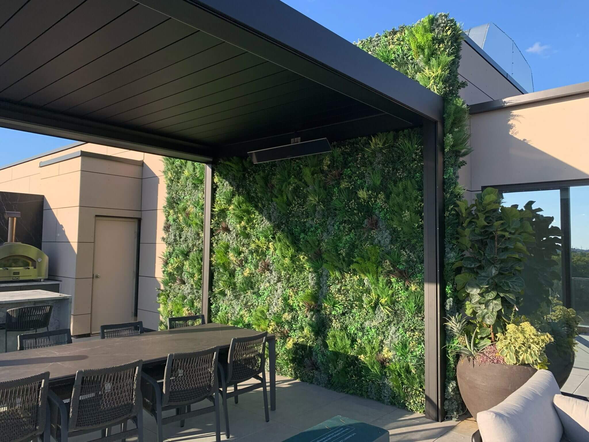 A modern roof terrace in Burlington, Ontario, features a dining table with chairs, a wood-fired oven, and a lush vertical garden wall under a pergola. The sunlit setting boasts a clear blue sky in the background.