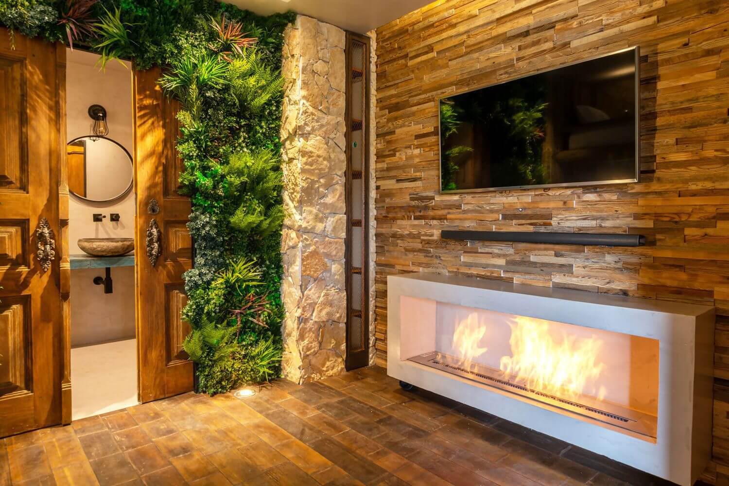 A cozy room features a modern fireplace with a vivid flame, a wooden accent wall with a mounted TV, and lush artificial plant walls that truly embrace nature at home. A stone wall section complements the view into a bathroom with a round mirror and wooden doors.