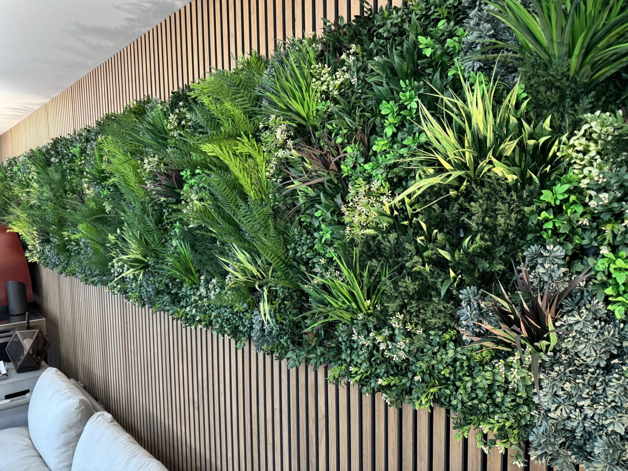 A vertical garden featuring a lush variety of green plants is mounted on a wooden slatted wall. A white sofa is positioned beneath the garden, enhancing the cozy, natural ambiance of the room.