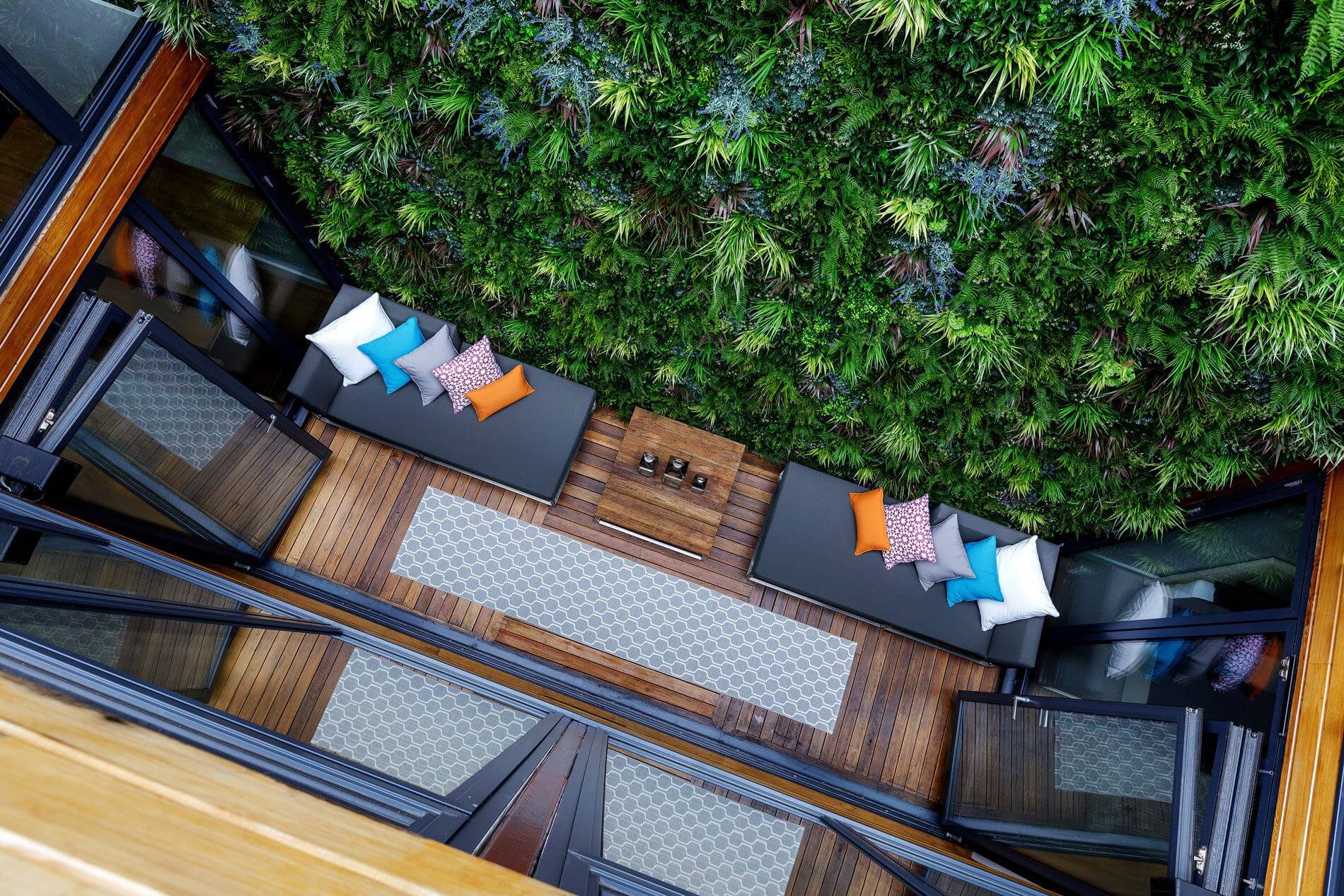 A cozy outdoor living space features two wooden benches lined with colorful cushions. An artificial vertical garden wall surrounds the area, enhancing the serene, nature-inspired atmosphere. A coffee table rests on a patterned rug between the benches.